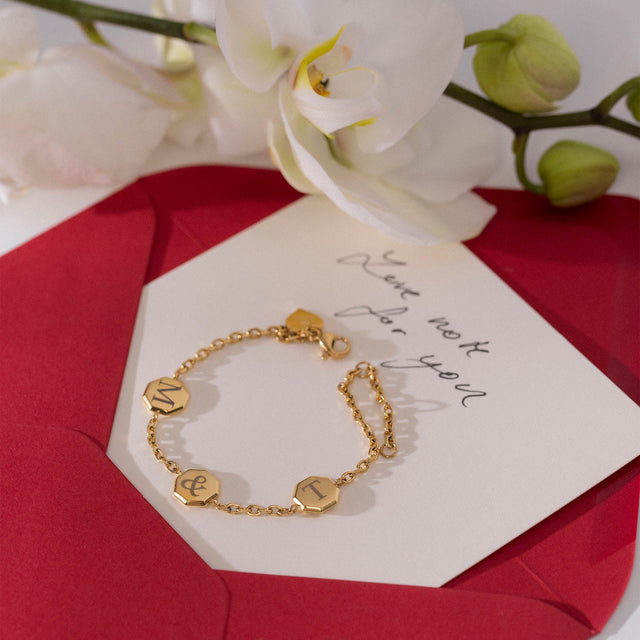 Goldenes Armband mit Buchstabenanhängern auf rotem Umschlag, PURELEI inspiriert.