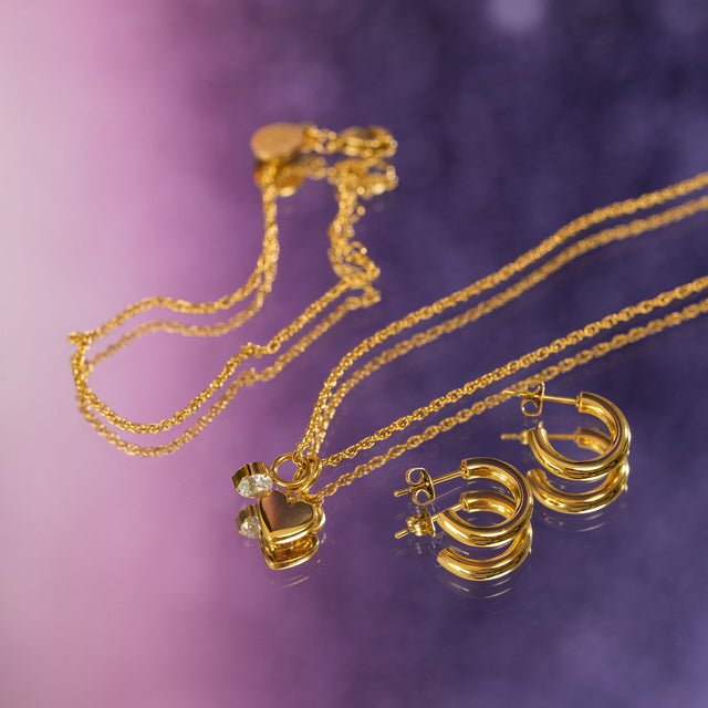 Goldene Kette mit Herz-Anhänger und Creolen von PURELEI auf lila Hintergrund.